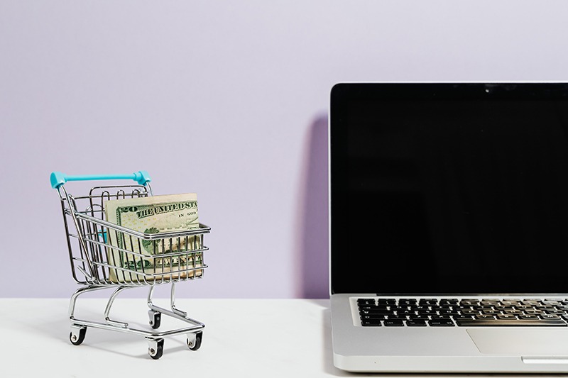 A small model of a shopping cart with money in it situated next to a laptop